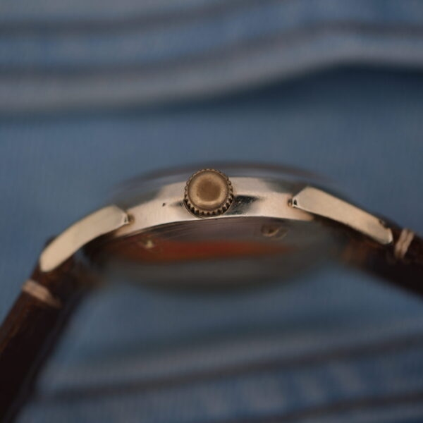 A close-up side view of the Longines Automatic Caliber 19AS wristwatch highlights its metal crown and vintage charm, complemented by a brown leather strap and softly blurred blue fabric background, evoking timeless elegance.