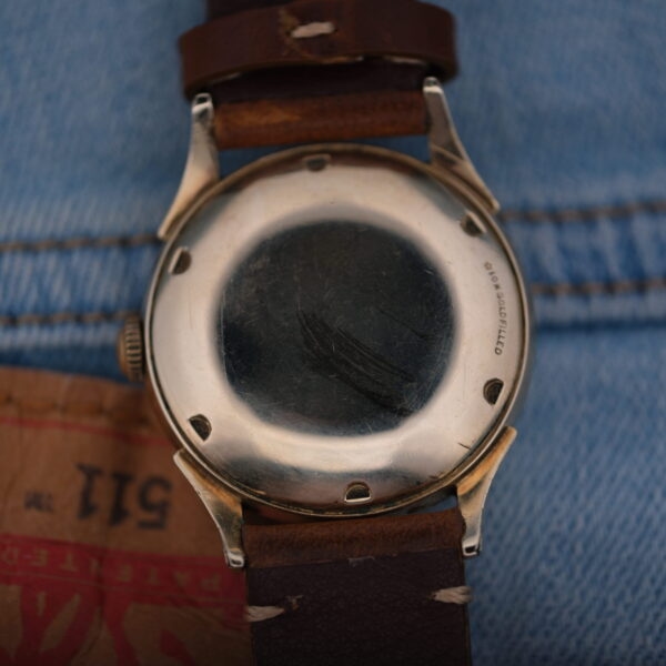A Longines Automatic Caliber 19AS watch with warm patina and twisted lugs, featuring a worn leather strap and scratched metal back, is displayed face down on denim fabric beside brown paper with partial red and black text.