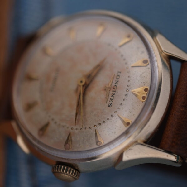 A close-up of the Longines Automatic Caliber 19AS Warm Patina & Twisted Lugs watch, featuring gold triangular markers and hands in a metal case, showcases timeless charm. This classic piece on a brown leather strap elegantly rests on a denim fabric background.