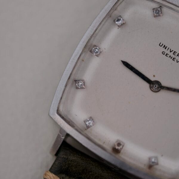 Close-up of the luxury Universal Geneve White Diamond Dial Micro-Rotor Cal. 66 wristwatch featuring a square silver case, diamond hour markers, black hands on a partially visible white dial with UNIVE... and GENÈVE text, complemented by a sleek black leather strap.