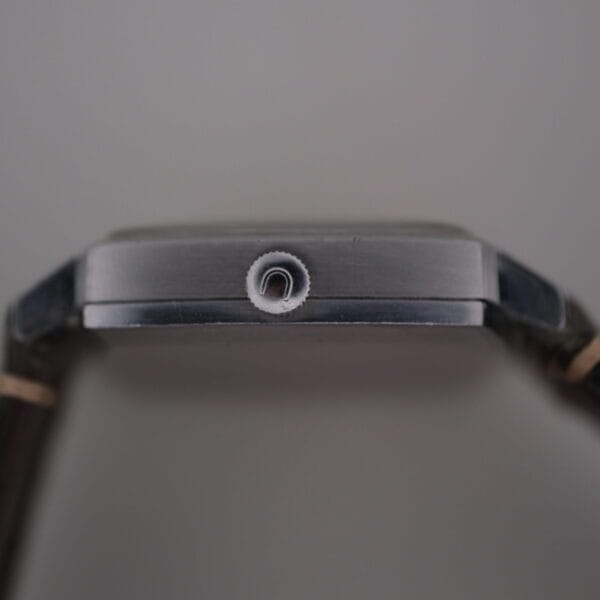 Close-up side view of a Universal Geneve White Diamond Dial Micro-Rotor Cal. 66 watch with a silver crown and dark wristband, showcasing its sleek metallic casing against a blurred backdrop that emphasizes the minimalist design.