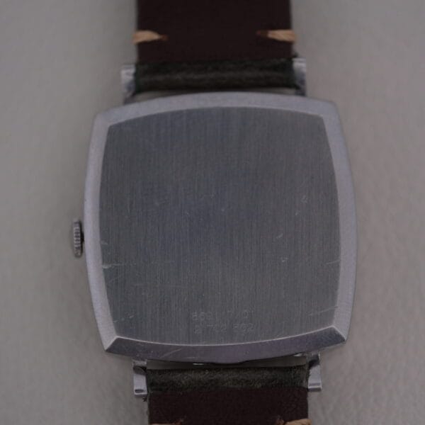 A close-up of a Universal Geneve White Diamond Dial Micro-Rotor Cal. 66 wristwatch reveals its brushed metallic finish. The dark leather strap, showing light wear and visible stitching, accompanies faint engravings near the bottom edge.
