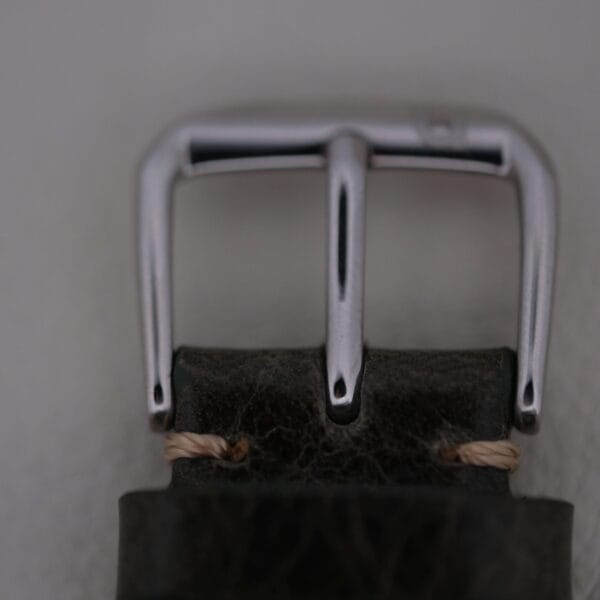 Close-up of a metallic Micro-Rotor buckle on a dark leather strap with light brown stitching. The focus highlights the shiny texture against a plain background. Based on Universal Geneve White Diamond Dial Micro-Rotor Cal. 66.