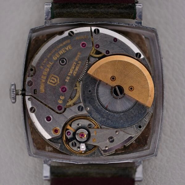 Close-up of a Universal Geneve White Diamond Dial Micro-Rotor Cal. 66 watch movement, showcasing intricate gears and parts. The open back reveals the internal mechanism with visible screws and inscriptions, with metal components featuring a stainless steel and brass finish, highlighting craftsmanship.