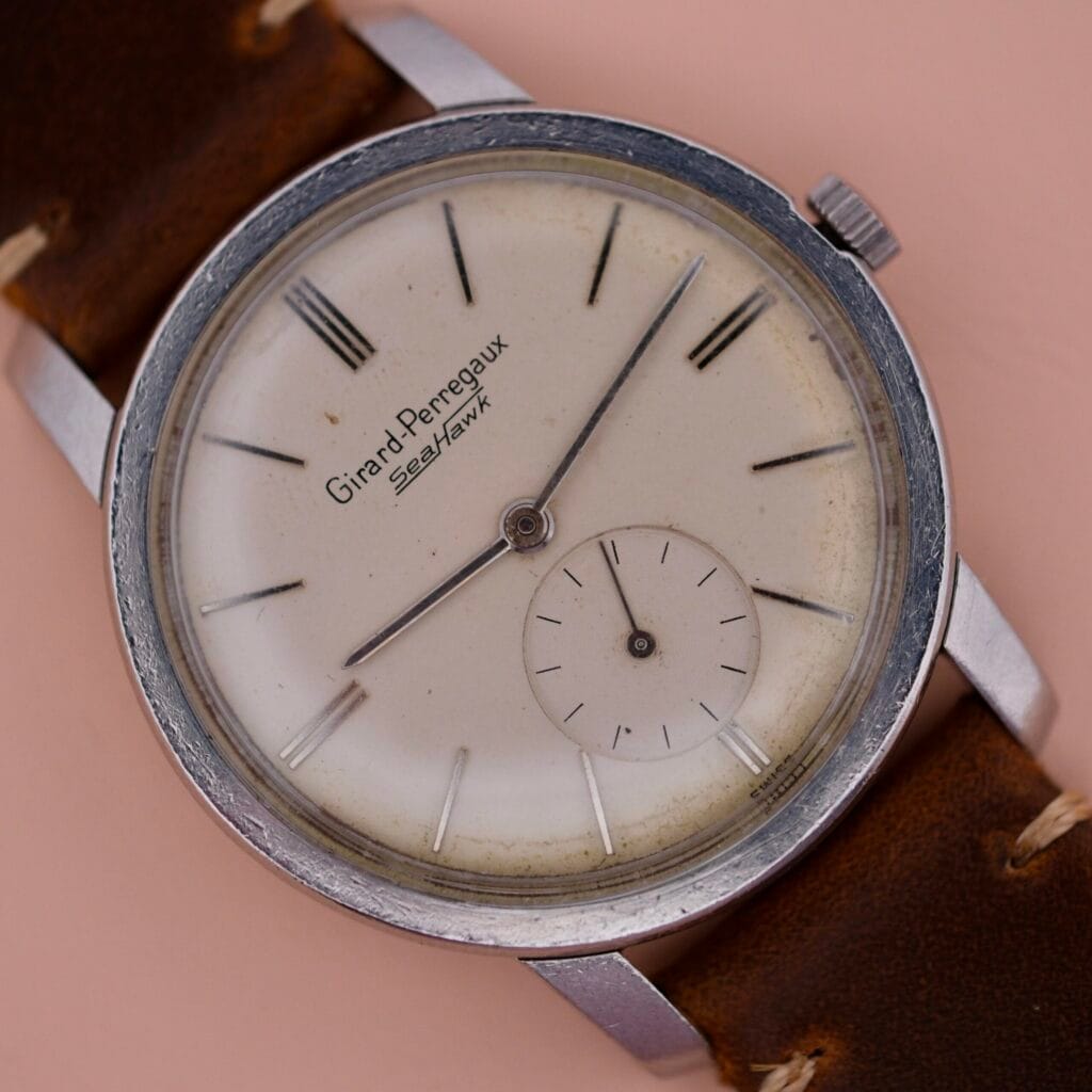 The vintage Girard Perregaux Sea Hawk wristwatch features a silver case, tan leather strap, and an aged white round dial with black hour markers and hands. A small sub-dial enhances its charm at the 6 oclock position.