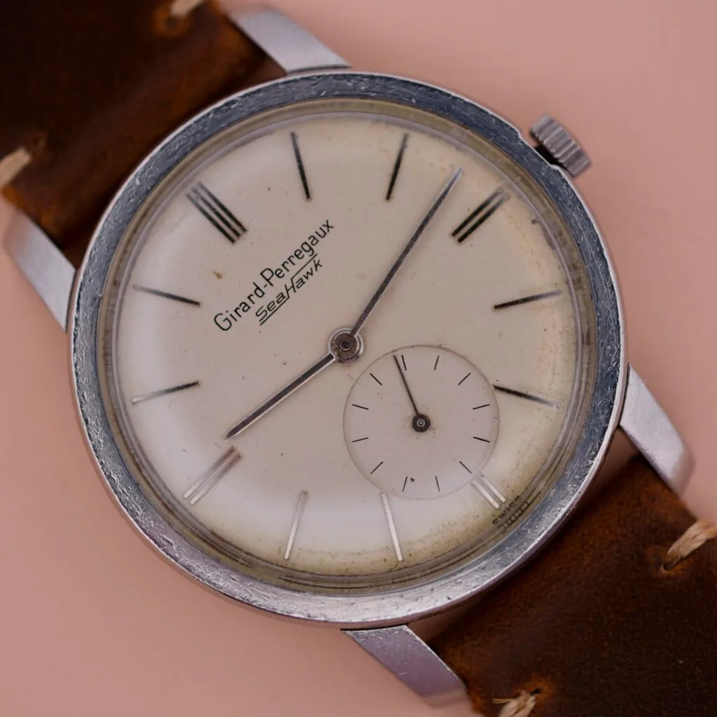 The vintage Girard Perregaux Sea Hawk wristwatch features a silver case, tan leather strap, and an aged white round dial with black hour markers and hands. A small sub-dial enhances its charm at the 6 oclock position.