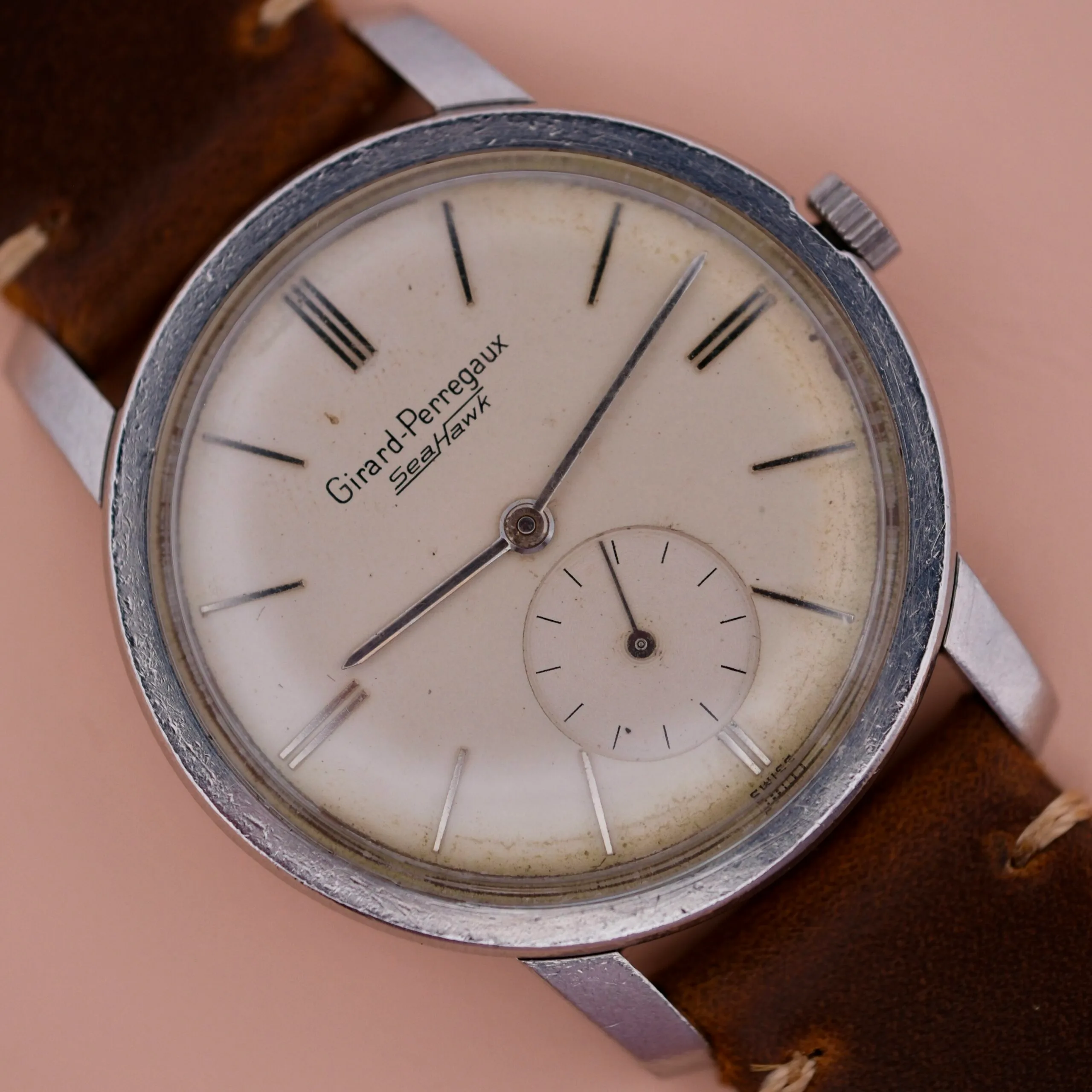 The vintage Girard Perregaux Sea Hawk wristwatch features a silver case, tan leather strap, and an aged white round dial with black hour markers and hands. A small sub-dial enhances its charm at the 6 oclock position.