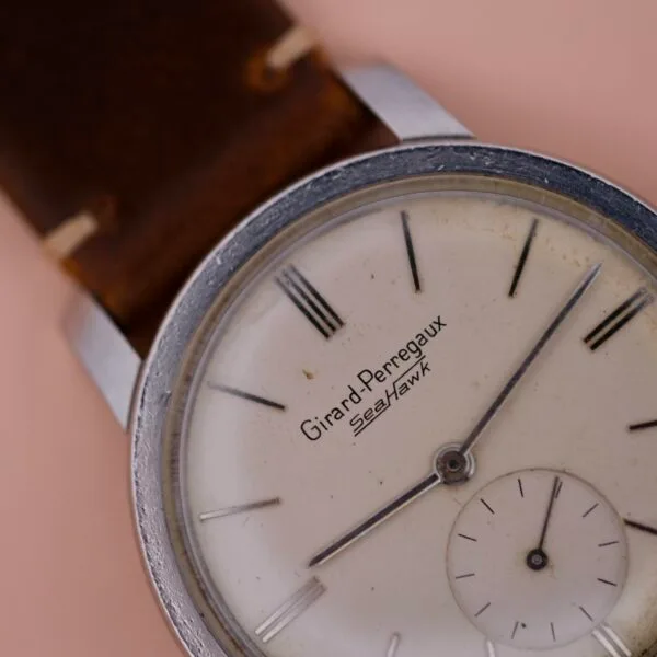 Close-up of the iconic Girard Perregaux Sea Hawk watch, highlighting its vintage allure with a silver round case and white dial. It boasts Roman numerals, minute markers, and a small subdial, enhanced by a brown leather strap in the backdrop.