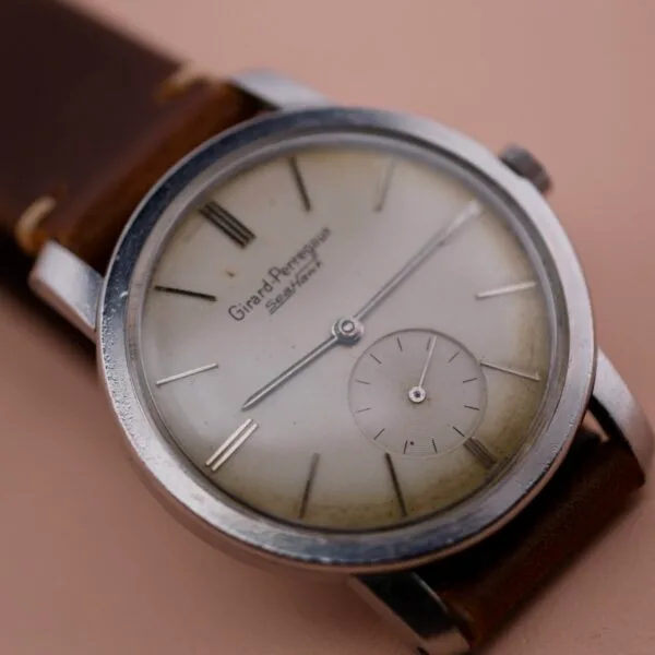 A vintage Girard Perregaux Sea Hawk wristwatch features a worn leather strap, an off-white round dial with simple hour markers, two hands, and a small seconds subdial. The slightly tarnished stainless steel case enhances its timeless charm.