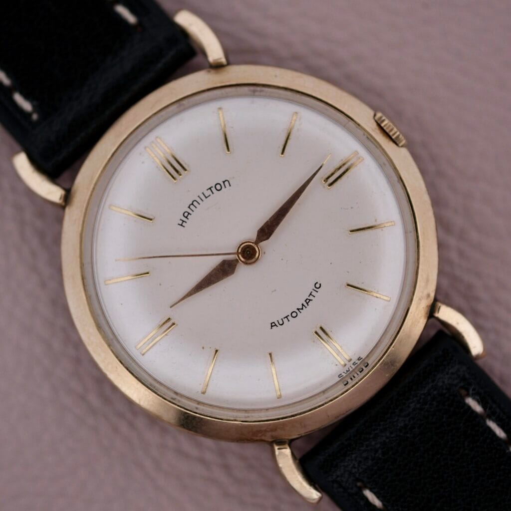 Close-up of a Hamilton Kinematic Swiss Automatic Fancy Lugs wristwatch with a white dial, gold case, and black leather strap.