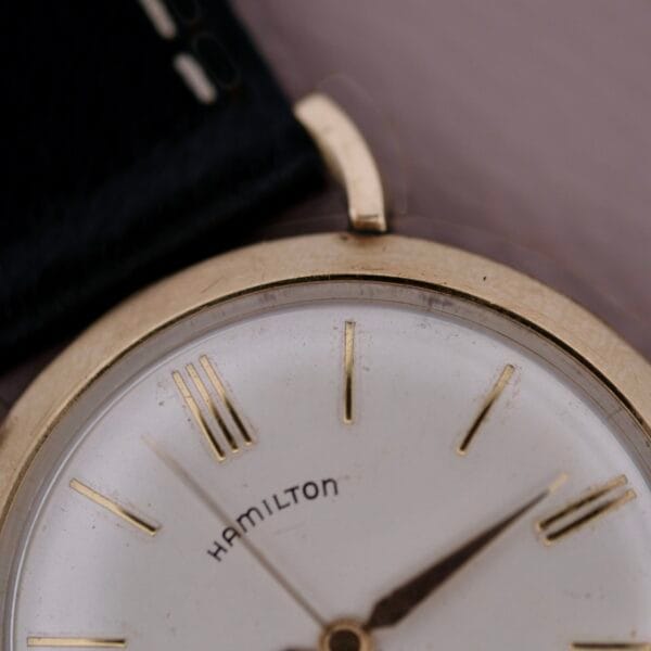 A close-up of the Hamilton Kinematic Swiss Automatic Fancy Lugs wristwatch showcases its vintage charm, with a classic white dial, elegant gold-tone hour markers and hands, and a glimpse of its sophisticated black leather strap completing the picture.