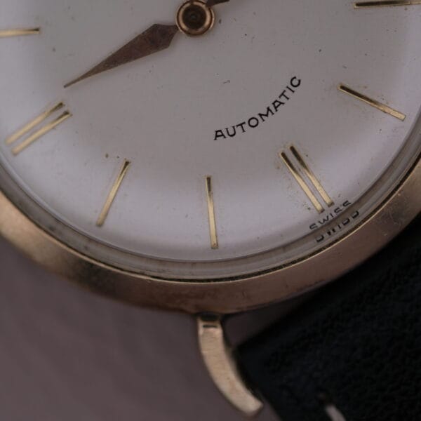 Close-up of a Hamilton Kinematic Swiss Automatic with white dial and gold markers. Automatic is featured on the face. The time reads approximately 10:08, and a black leather strap is partially visible.