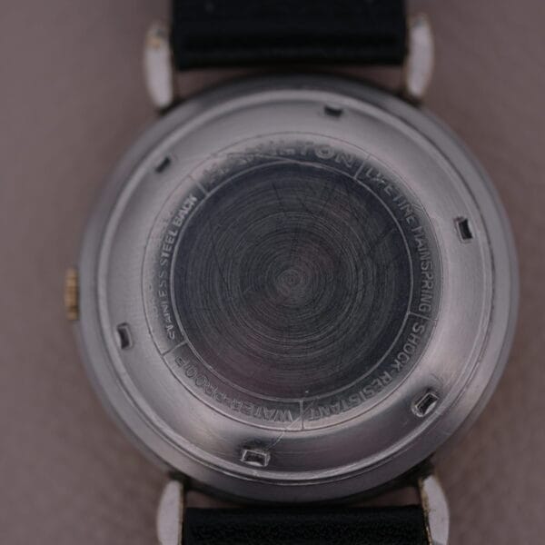 The close-up reveals the back of the Hamilton Kinematic Swiss Automatic Fancy Lugs watch, featuring engravings such as Hamilton, Stainless Steel Back, Shock Resistant, and Waterproof. A sleek black strap is securely attached.