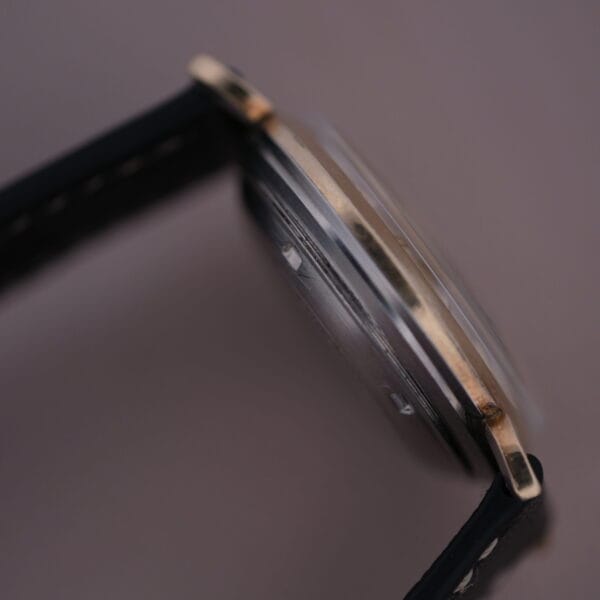 Close-up of the Hamilton Kinematic Swiss Automatic Fancy Lugs watch, highlighting its slim profile. This wristwatch has a black leather strap with a gold-toned bezel, exuding minimalistic elegance against a neutral background.