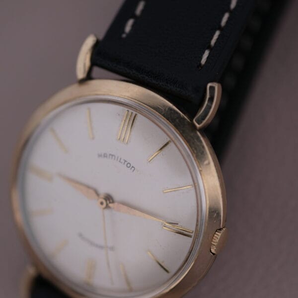 A close-up of the Hamilton Kinematic Swiss Automatic Fancy Lugs watch highlights its vintage charm with a gold case and white dial. Featuring thin gold hour markers, hands, and the Hamilton logo, it is complemented by a black leather strap with white stitching, exuding timeless elegance.