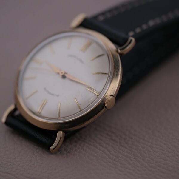 A close-up of the Hamilton Kinematic Swiss Automatic Fancy Lugs wristwatch shows its gold case, white dial, and black leather strap. Its minimalist hour markers and three hands exude timeless elegance against a textured surface background.
