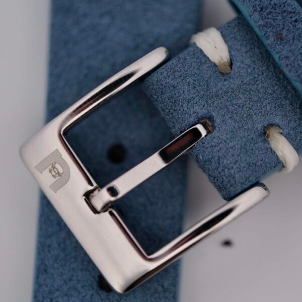 Close-up of the silver watch buckle with engraved emblem, attached to a Powder Blue Suede Watch Strap Vintage Stitch featuring white stitching.