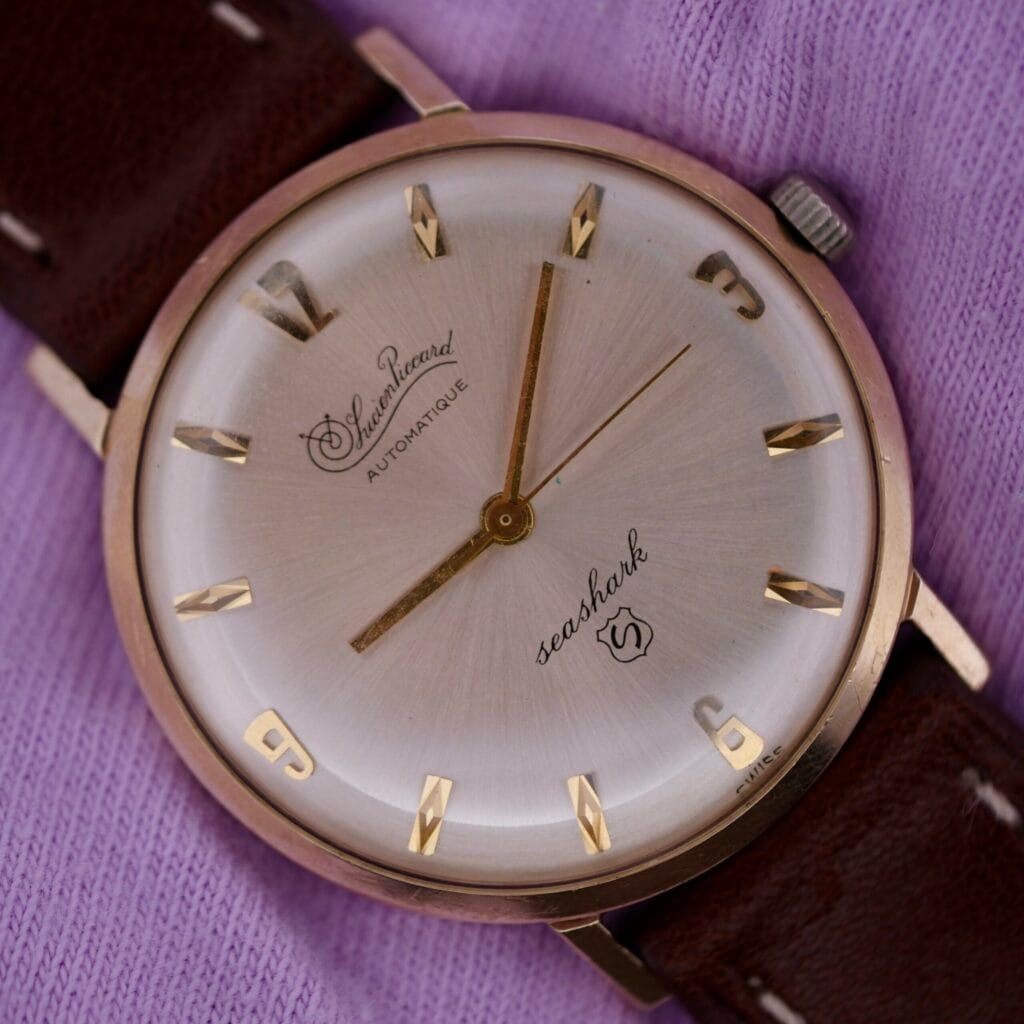 The Lucien Picard Seashark Automatic Slim Case is a vintage wristwatch featuring a brown leather strap, round silver face, gold hour markers, and elegant hands, all displayed gracefully against a purple fabric background.