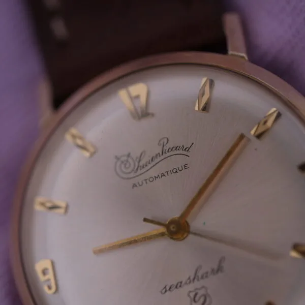Close-up of a vintage wristwatch with a brown leather strap, cream face, and gold hour markers and hands. Labeled Lucien Picard Seashark Automatic Slim Case, it rests on soft purple fabric, reminiscent of classic designs.