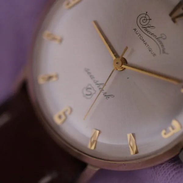 Close-up of a vintage Lucien Picard Seashark Automatic Slim Case wristwatch with gold hour markers and hands against a brown leather strap, set against a softly blurred purple background.