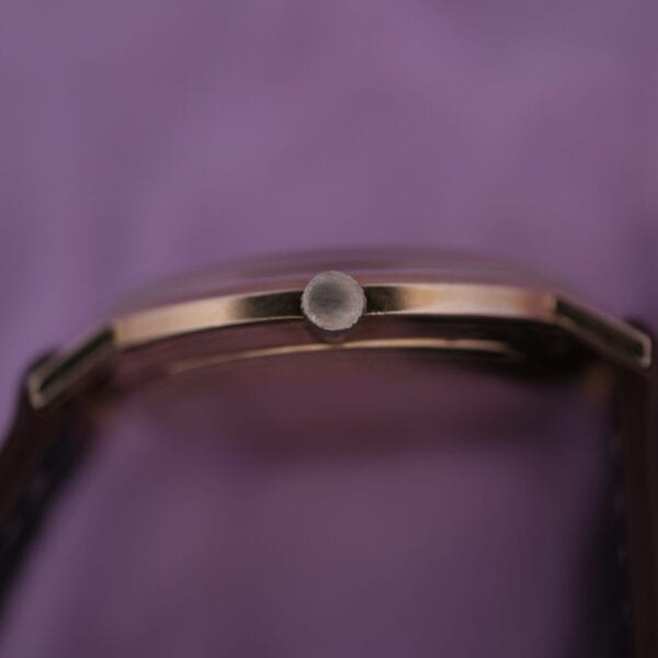 Close-up of the Lucien Picard Seashark Automatic Slim Cases side, highlighting the crown with a blurred purple backdrop. It showcases a gold-toned body and sleek black strap.