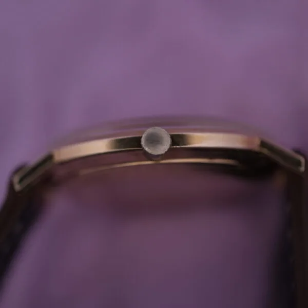 Close-up of the Lucien Picard Seashark Automatic Slim Cases side, highlighting the crown with a blurred purple backdrop. It showcases a gold-toned body and sleek black strap.