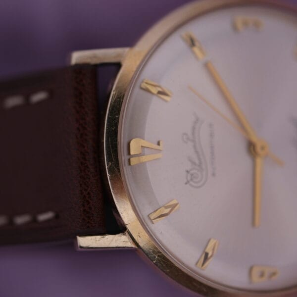 Close-up of a Lucien Picard Seashark Automatic Slim Case watch with a gold bezel, white face, and gold hour markers and hands, on a brown leather strap against a soft purple fabric background.