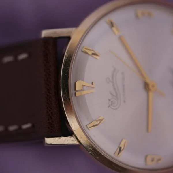Close-up of a Lucien Picard Seashark Automatic Slim Case watch with a gold bezel, white face, and gold hour markers and hands, on a brown leather strap against a soft purple fabric background.