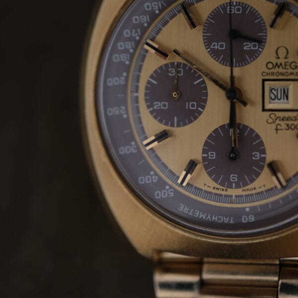 Close-up of a 1975 Omega Speedsonic f 300Hz Chronograph Ref. 188.0002 watch face, featuring three sub-dials and a date window on a dark background, with Tachymètre engraved on the bezel. It has a brass-colored case and band with intricate markings.