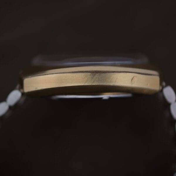 Close-up side view of a 1975 Omega Speedsonic f 300Hz Chronograph Ref. 188.0002 with a gold-colored case and glass cover. The visible scratches and small dent add character. The watch is attached to a metal bracelet, set against a dark background.