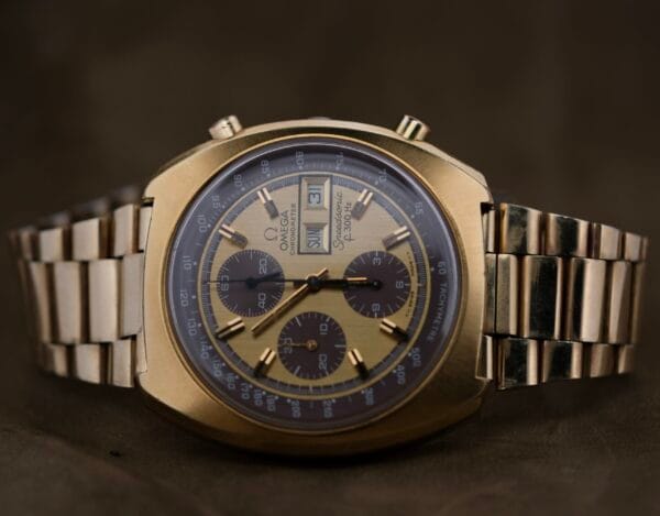 The 1975 Omega Speedsonic f 300Hz Chronograph Ref. 188.0002 features a gold detailed face with three sub-dials and a date window, paired with a metallic bracelet, set elegantly against a dark background.