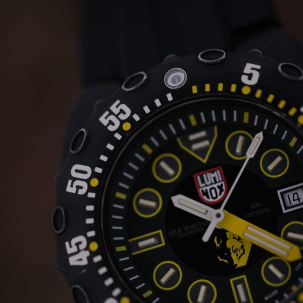 A close-up of the Luminox Scott Cassell Deep Dive Special 1526 Automatic shows a black analog watch with yellow accents, Sea Wolves and 500 meters text. It features white hour markers, a date display, signature design elements, white numerals on the bezel, and a black strap.