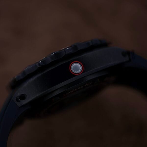 Close-up of a black Luminox Scott Cassell Deep Dive Special 1526 Automatic wristwatch side with a round button featuring a red outline, showcasing its rugged design; the textured bezel is visible on top against a blurred dark brown background.