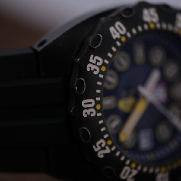 Close-up of a black Luminox Scott Cassell Deep Dive Special 1526 Automatic watch, showcasing a detailed bezel with bold white numerals on black. The navy blue dial has yellow accents and white hour markers, with the focus on the bezel and a slightly blurred dial.