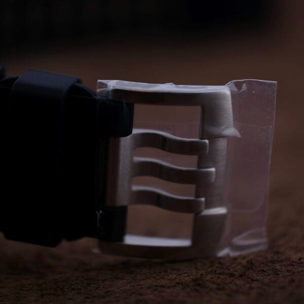 Close-up of a Luminox Scott Cassell Deep Dive Special 1526 Automatic watch buckle, partially wrapped in protective plastic, attached to a black strap, resting on a brown surface.