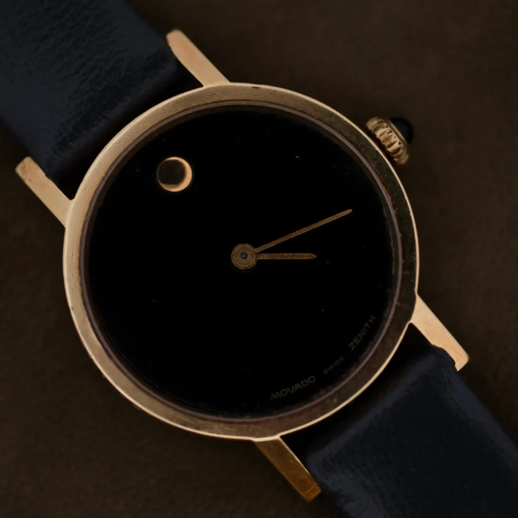A Ladies Movado Zenith 14k Gold Museum Dial wristwatch with a black face and leather strap rests elegantly on a brown surface.