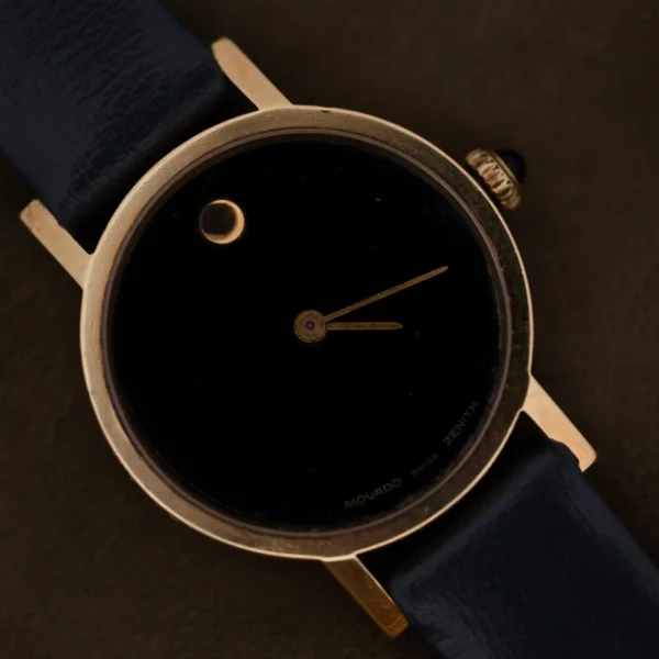 A close-up of a Ladies Movado Zenith 14k Gold Museum Dial wristwatch with a minimalistic design, featuring a black dial with a single dot at 12 oclock, gold-tone hands, and an elegant black leather strap, exuding sophistication against the dark background.