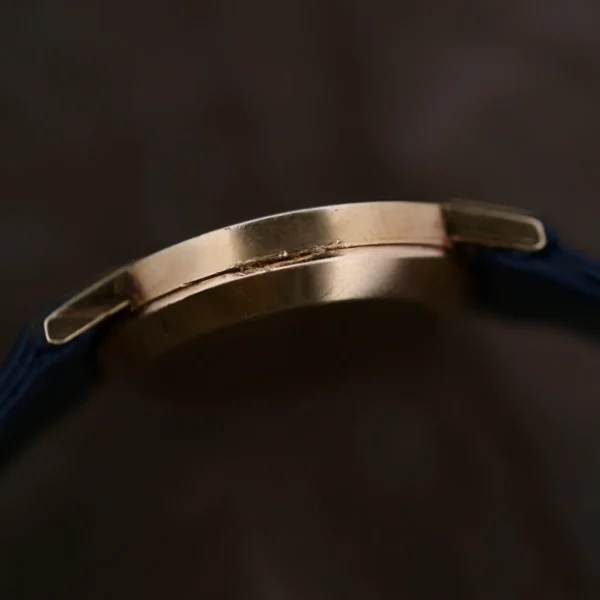 Side view of the Ladies Movado Zenith 14k Gold Museum Dial watch with a gold case and dark blue leather strap, elegantly highlighted by its slim profile against a dark, blurred backdrop.