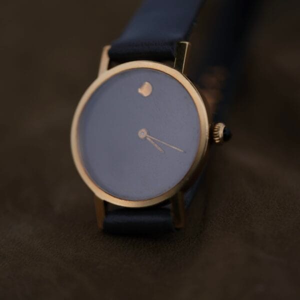 The Ladies Movado Zenith 14k Gold Museum Dial is a minimalist watch with a blank face inspired by Movados iconic design, featuring a gold-toned case, small side crown, and black leather strap against a dark textured background.