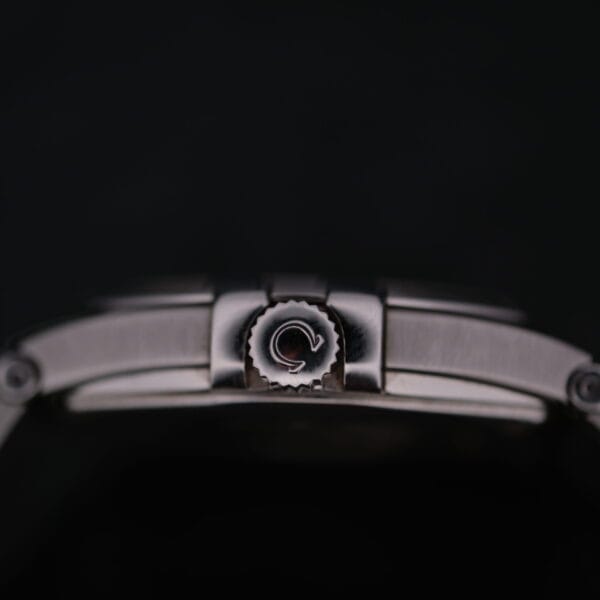 A close-up of the Omega Constellation Ref. 396.201 crown, featuring a logo, set against a dark backdrop highlights the intricate stainless steel details and rotating mechanism of this watchs side.