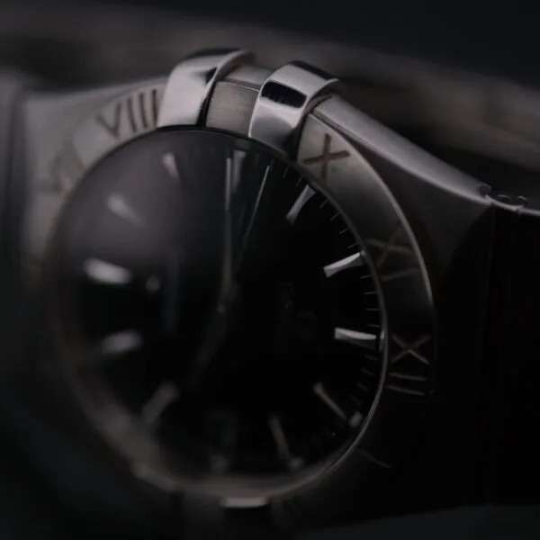 A close-up of the Omega Constellation Ref. 396.201 highlights its dark face with Roman numerals on the bezel, contrasted by a polished metallic case and band against a blurred backdrop, showcasing its sophisticated design and craftsmanship.