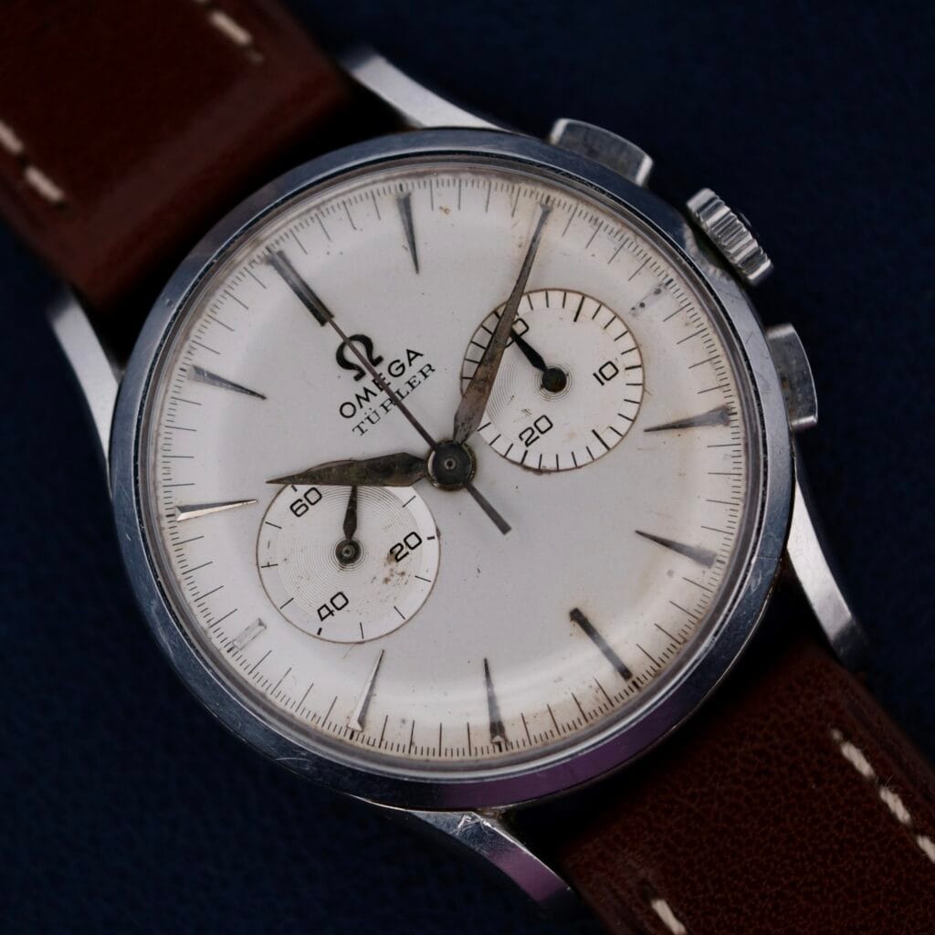 Omega Turler Dial Chronograph 2463/1 Caliber 320 featuring a white dial, two subdials, and a brown leather strap shown on a dark background.
