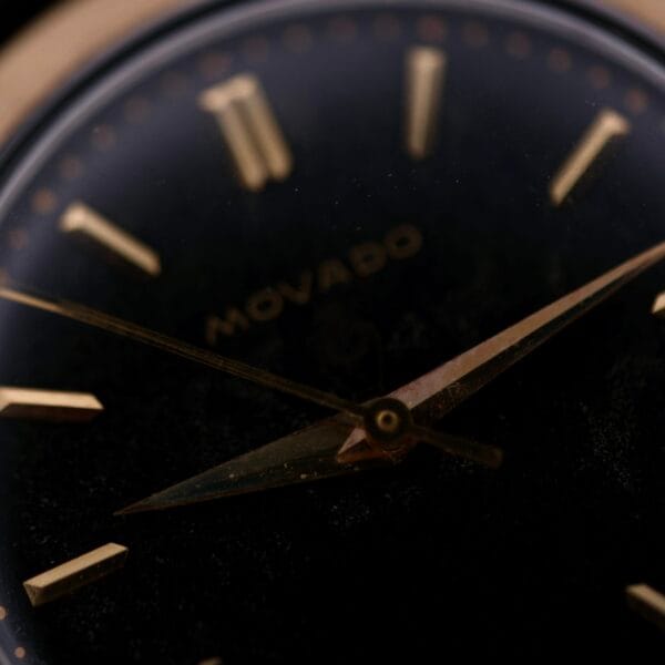 Close-up of a vintage Movado Ref. 18240 Francois Borgel Gold Top watch face, positioned against a black background, with gold hour markers. The hands, slightly aged, are prominently displayed and the Movado name is elegantly visible on the dial for the Hong Kong Market edition.