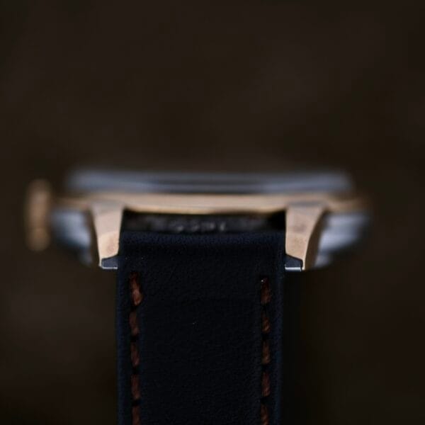 Close-up of a wristwatch viewed from the side, highlighting the elegant metal case of the Movado Ref. 18240 Francois Borgel Gold Top Hong Kong Market and a black leather strap with brown stitching set against a dark background, focusing on the strap while leaving the watch face blurred.