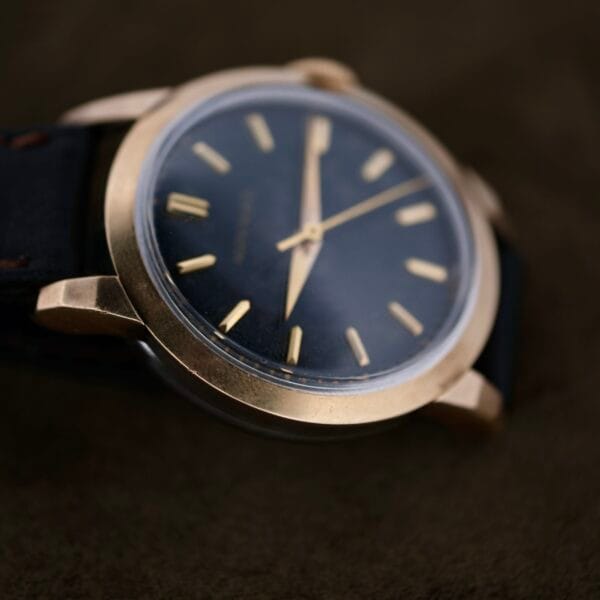 A close-up of a Movado Ref. 18240 Francois Borgel Gold Top showcases a vintage wristwatch with a gold-colored case and black leather strap. The watch features a black face with gold hour markers and hands, resting on a dark textured surface.