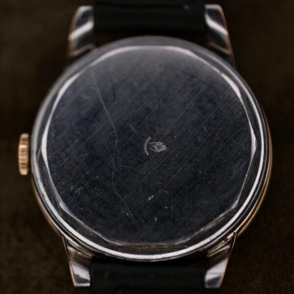 A close-up of the rear of a vintage Movado Ref. 18240 Francois Borgel Gold Top Hong Kong Market wristwatch reveals a scratched and worn metal cover with the faint outline of a small emblem or logo, while part of its black strap is visible.