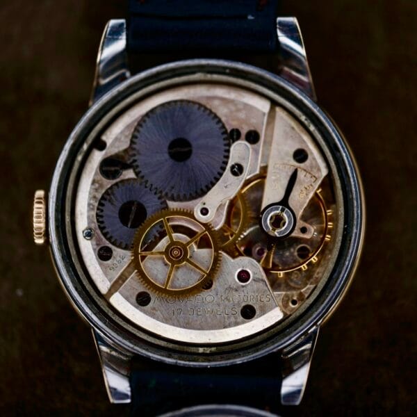 Close-up of a Movado Ref. 18240 Francois Borgel Gold Top mechanical watch, featuring an open back that reveals its intricate gears and components with 17 jewels on display. The metal parts interlock to showcase the craftsmanship and engineering, complemented by a black strap.