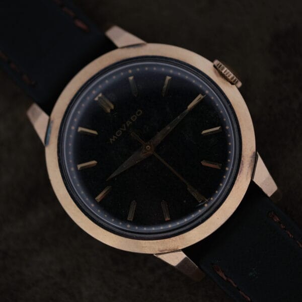 Close-up of the Movado Ref. 18240 Francois Borgel wristwatch with a gold top and black leather strap. This elegant Hong Kong Market timepiece has a minimalist design, featuring hour markers and two hands on a black dial against a dark background.