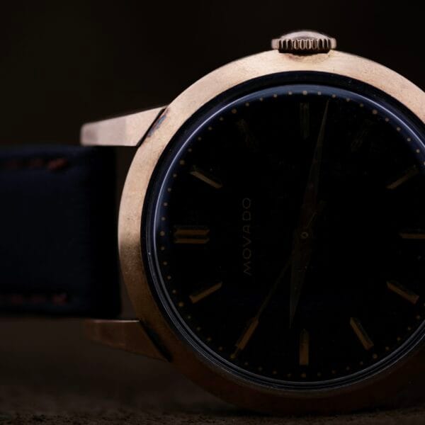 Close-up of a vintage Movado Ref. 18240 Francois Borgel wristwatch, featuring a dark face and gold casing. The hands and hour markers appear under dim lighting, with Movado subtly visible on the dial, complementing its elegant black strap.