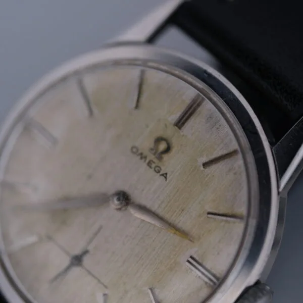 A close-up of the Omega Manual Wind Cal. 268 showcases its aged charm, featuring a beige dial with slender hour and minute markers and dark hands. The partially visible black strap enhances the timeless elegance of this classic timepiece.